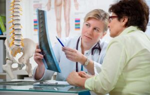 A person reviews a lower-back spine model with a clinician in a bright examination room.